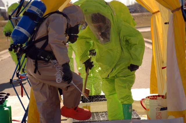 Two hazmat-suited responders decontaminating at a roadside station under a yellow tent, one in lime green and the other in beige, using brushes and hoses.